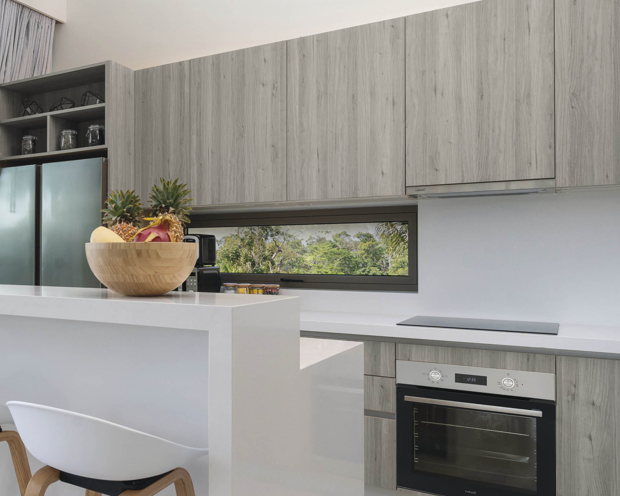 Cocina moderna con armarios de madera gris, isla blanca, frutero, horno empotrado y vista a la vegetación.