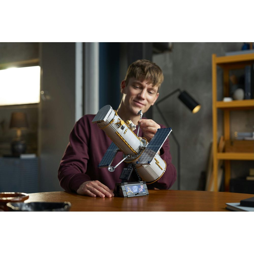 Un joven sentado en una mesa de madera construye un modelo detallado de Lego del telescopio espacial Hubble.