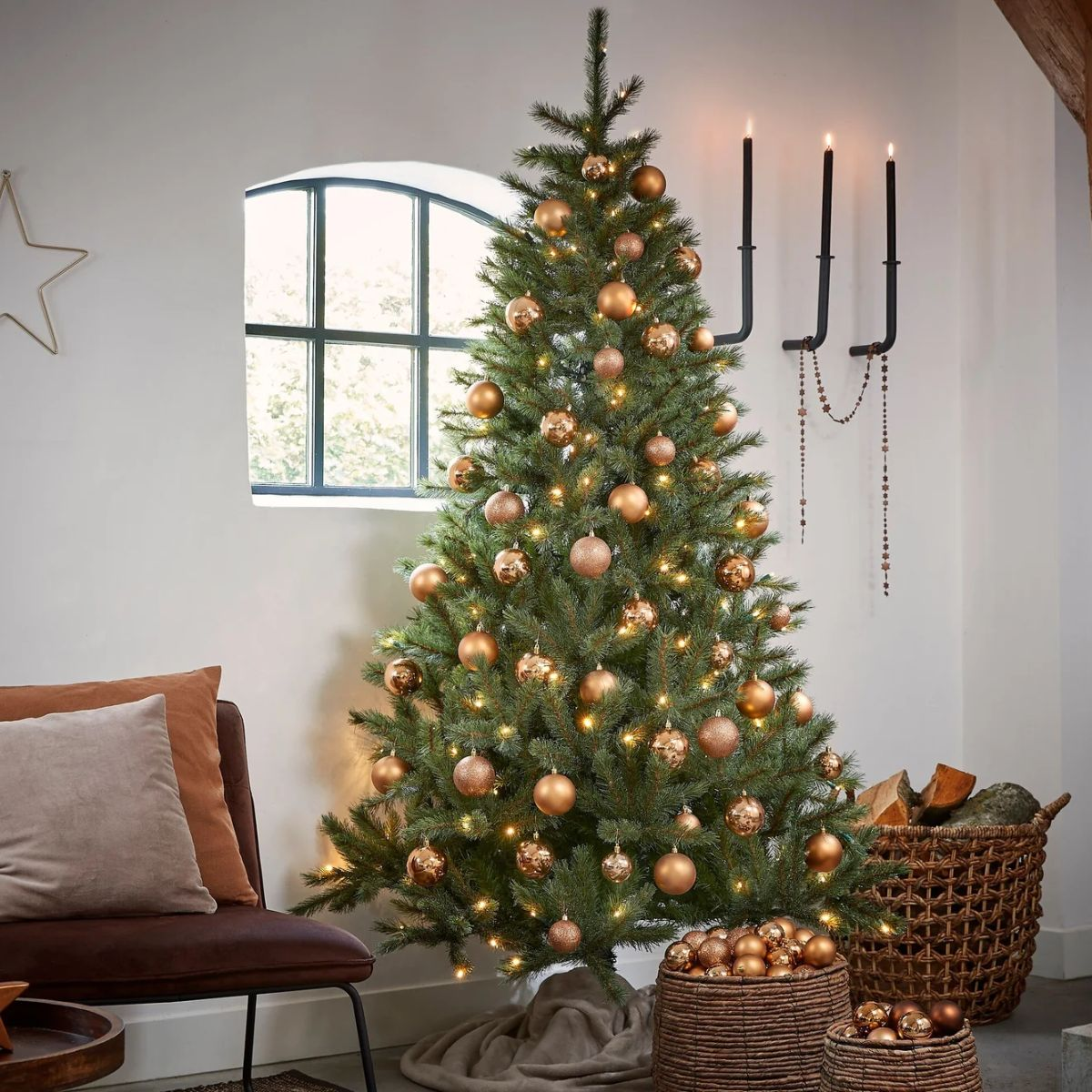 Een versierde kerstboom met gouden ballen en lampjes in een gezellige kamer met fauteuils en haardhout.