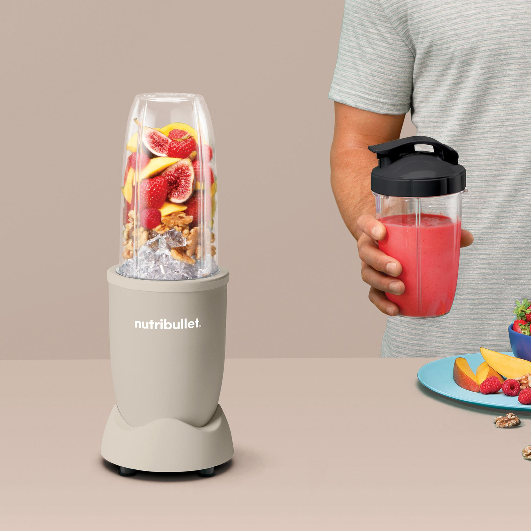 A Nutribullet blender filled with fruit and ice stands on a table as a person holds a red smoothie cup.