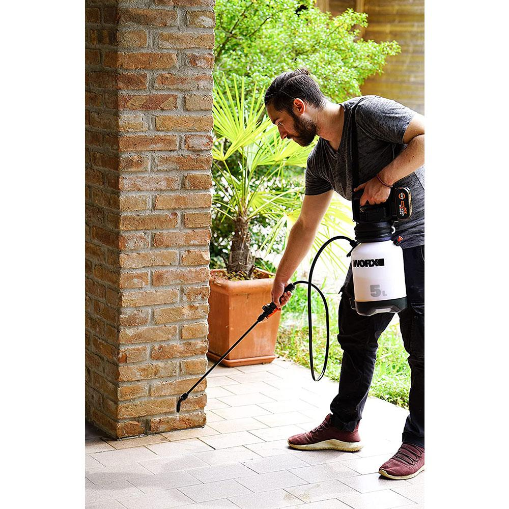 Bir adam fayanslı bir verandada Worx basınçlı püskürtücü ile tuğla duvarı suluyor.