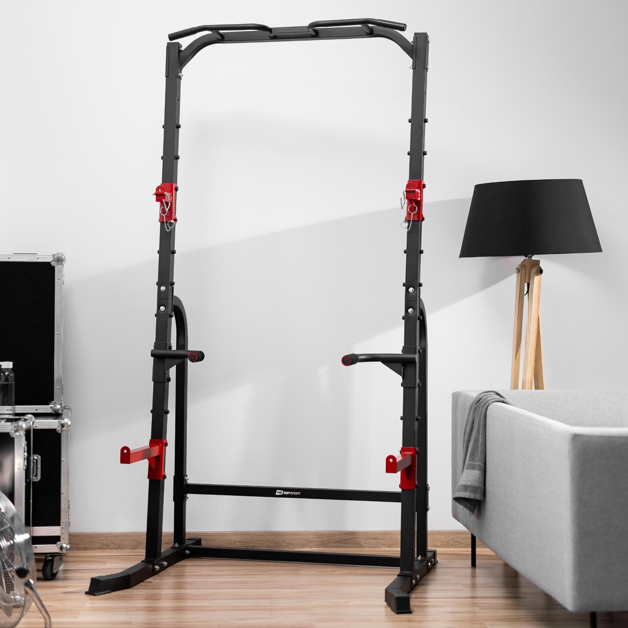 Ein schwarz-rotes Metall-Power-Rack steht in einem Raum mit grauem Sofa, Holzlampe und gestapelten Koffern im Hintergrund.