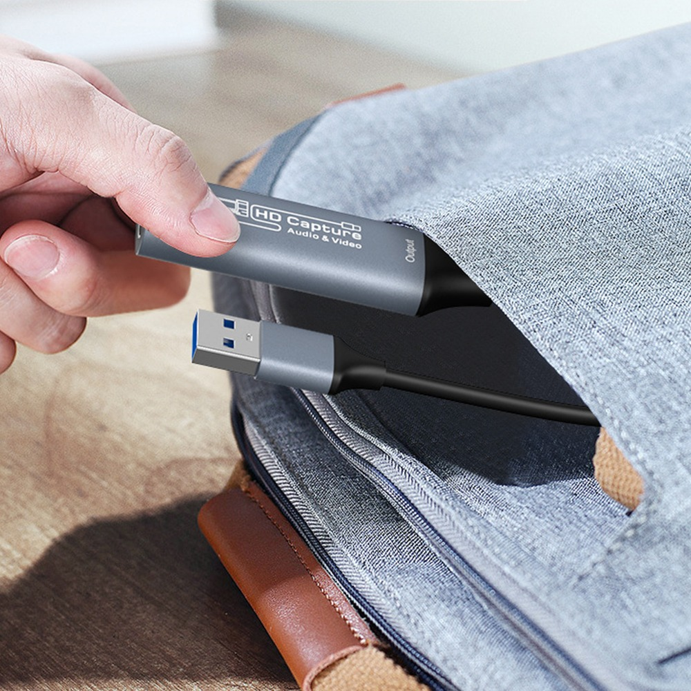 Eine Hand legt ein HD-Capture-Gerät mit USB-Kabel in eine graue Tasche.