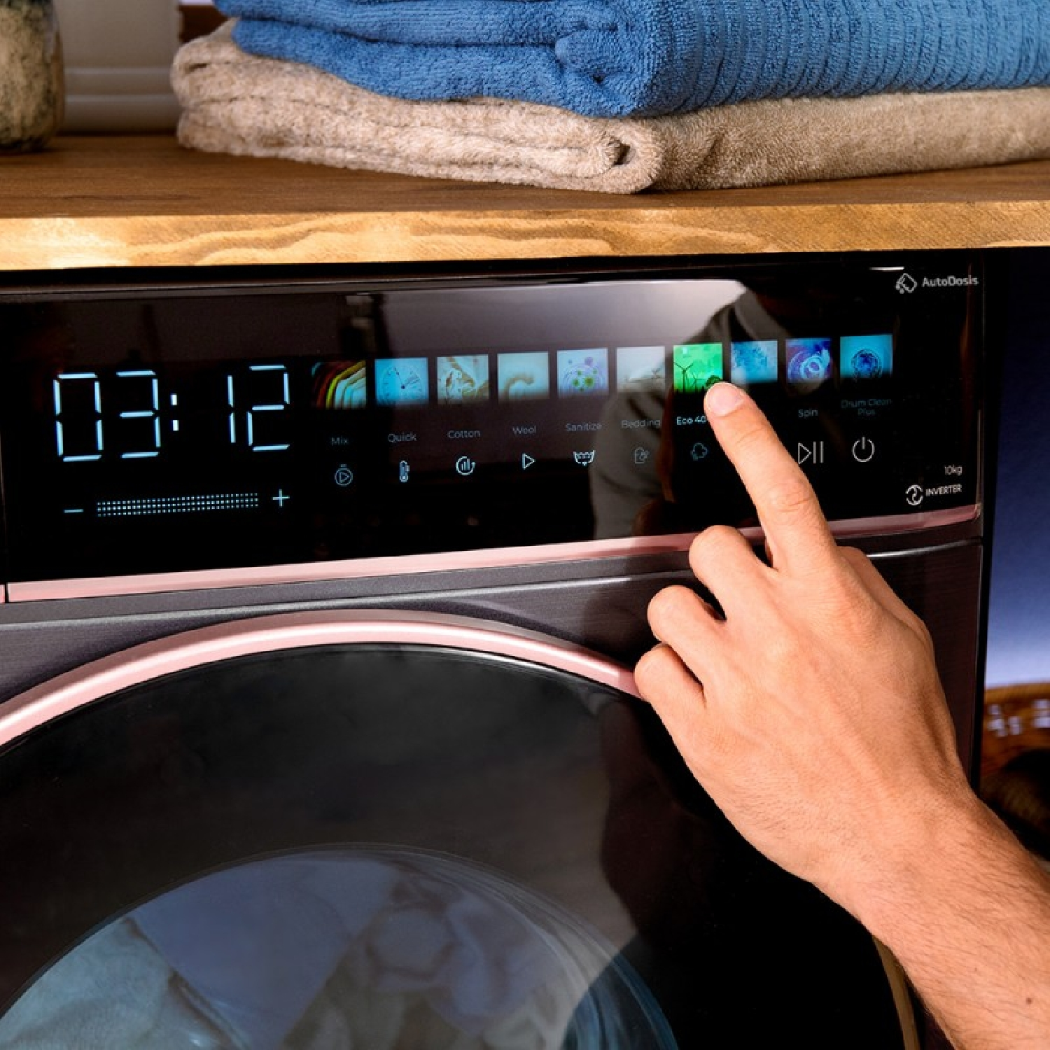 Una mano interactúa con el panel de control digital de una lavadora moderna con toallas dobladas.