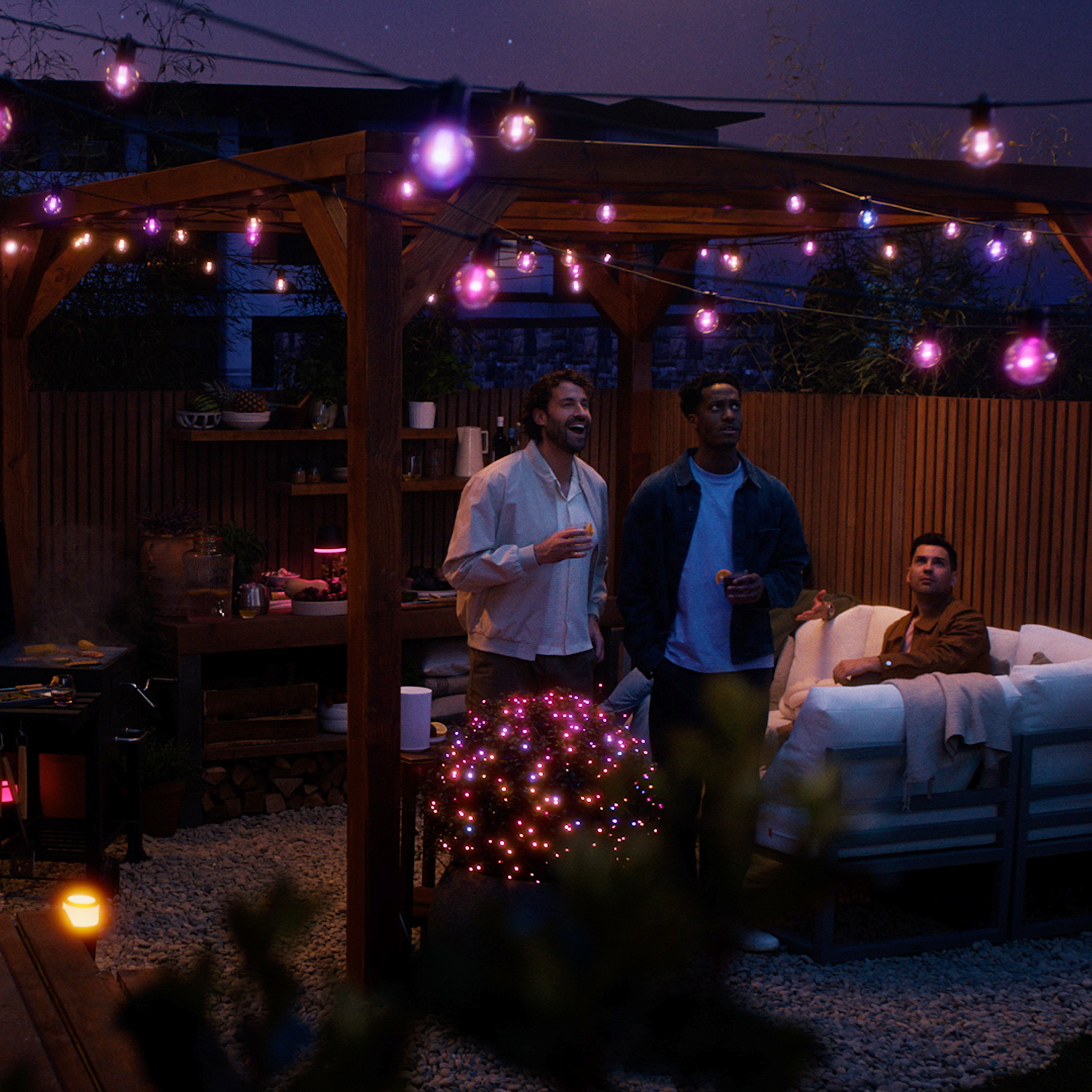 Trois hommes discutent sur un patio au crépuscule, éclairés par des guirlandes lumineuses violettes.