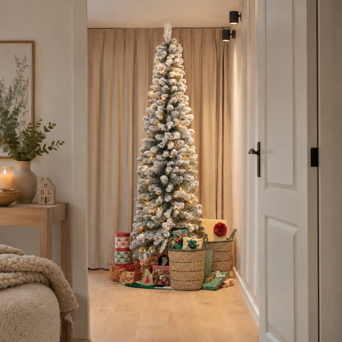 A slim, snow-flocked Christmas tree with lights is set in a room with wrapped gifts in baskets at its base.