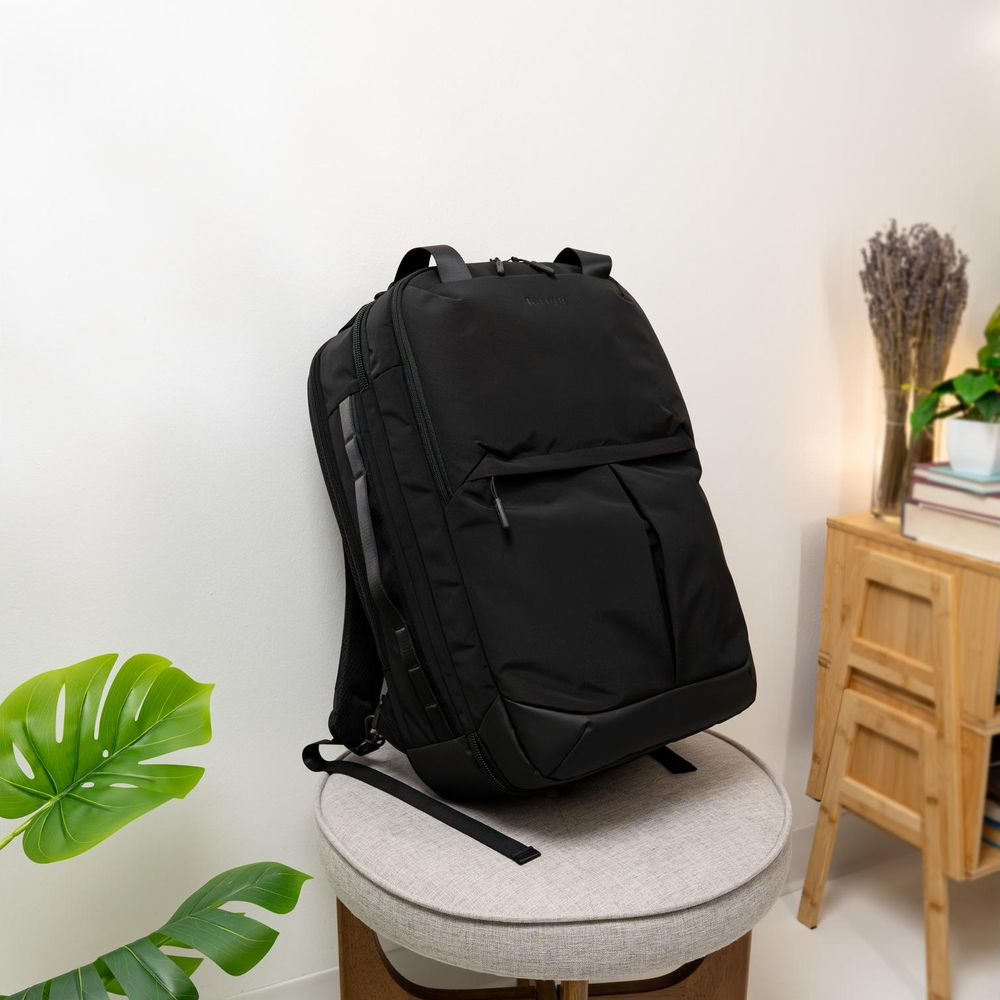 Ein schwarzer Rucksack steht auf einem runden, grauen Hocker in einem Raum mit weißen Wänden und Pflanzen.