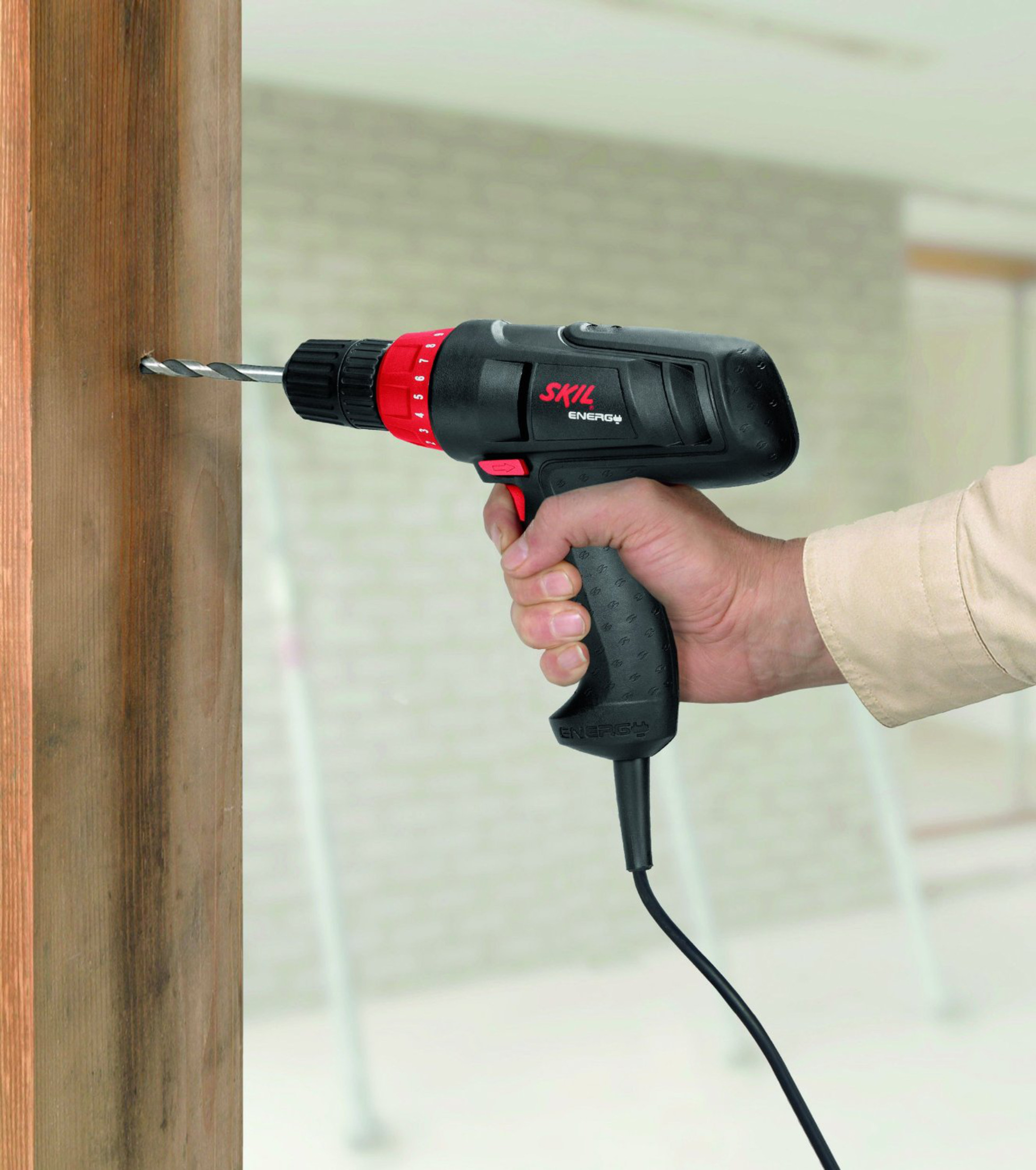 Una mano fora una trave di legno usando un trapano elettrico Skil nero e rosso.