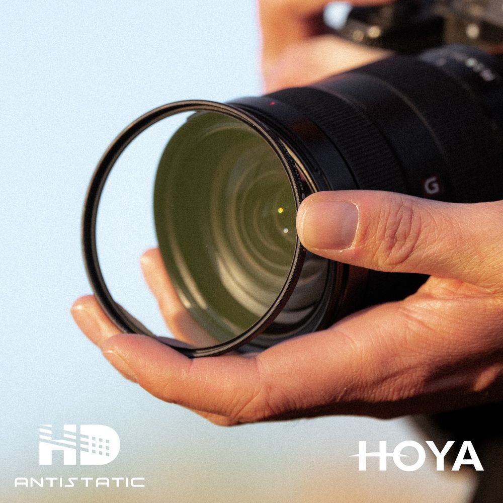 Eine Hand hält einen Kamerafilter vor ein Kameraobjektiv mit der Aufschrift HOYA ANTISTATIC.