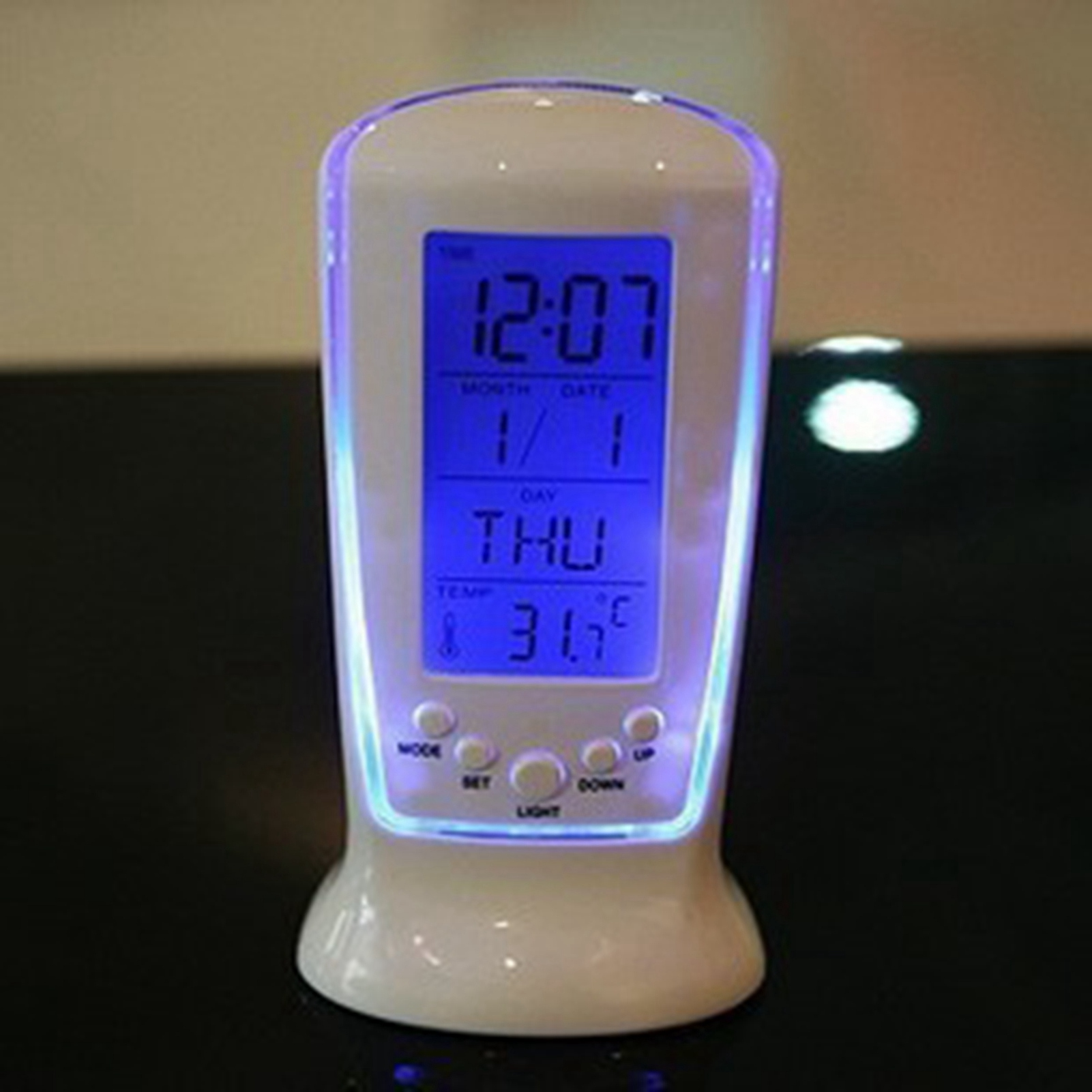 Ein weißer digitaler Wecker mit blauem Display, Uhrzeit, Datum und Temperatur auf einem dunklen Tisch.