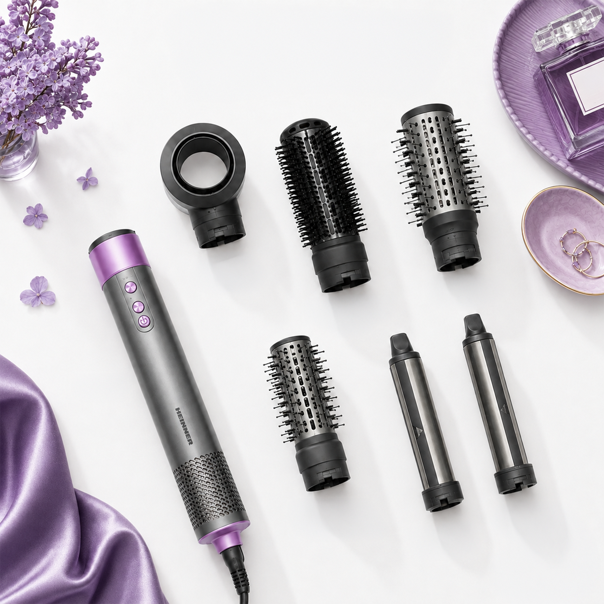 Appareil coiffant gris avec accessoires sur fond blanc, entouré de fleurs violettes et d'un tissu en soie.