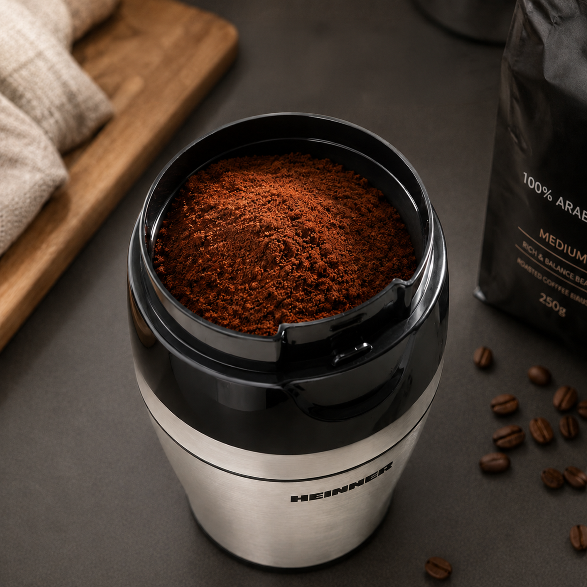 Eine Kaffeemühle aus Edelstahl mit frisch gemahlenem Kaffeepulver, umgeben von Kaffeebohnen und einer Kaffeetüte.