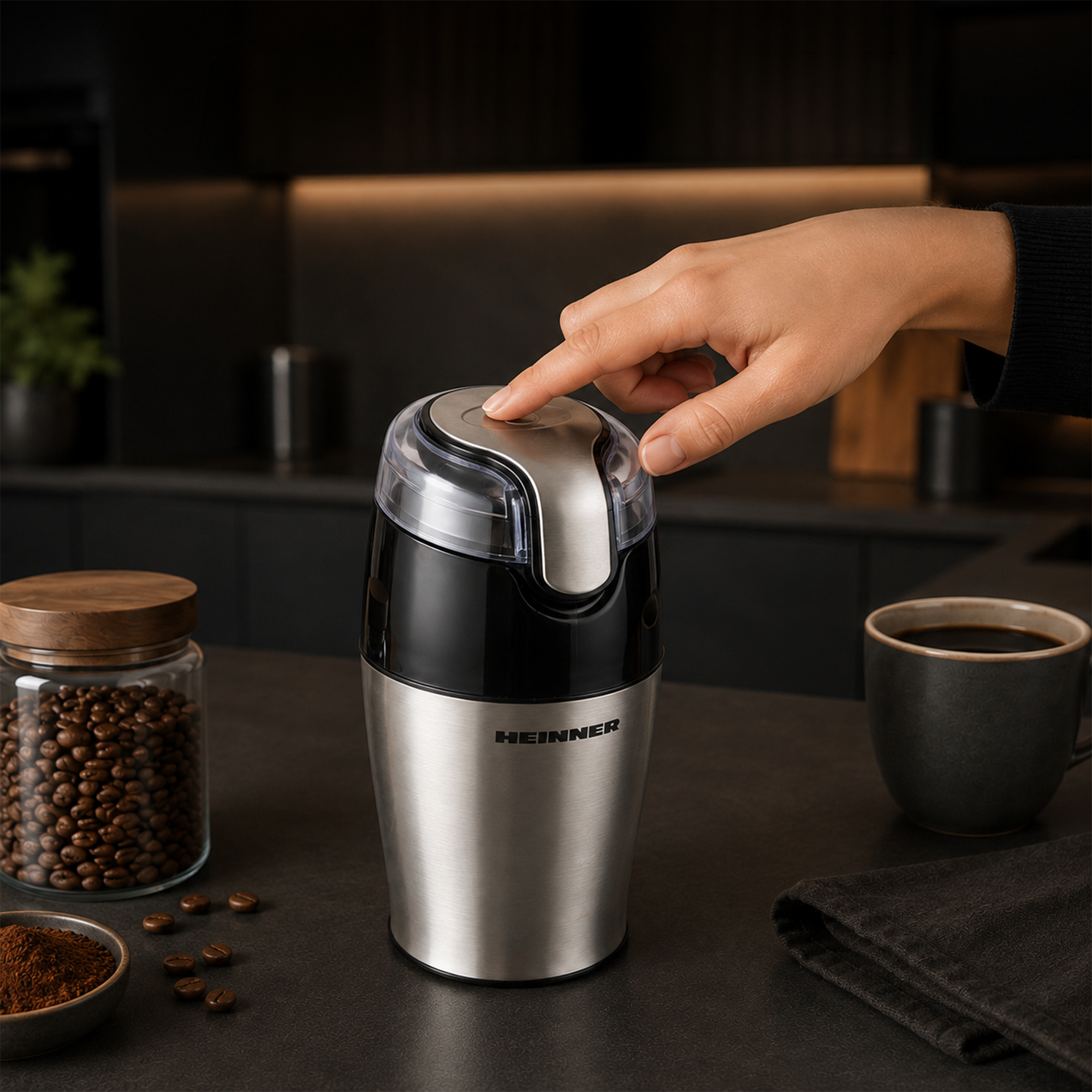 Eine Hand drückt den Startknopf einer silber-schwarzen Heinner Kaffeemühle in einer Küche.
