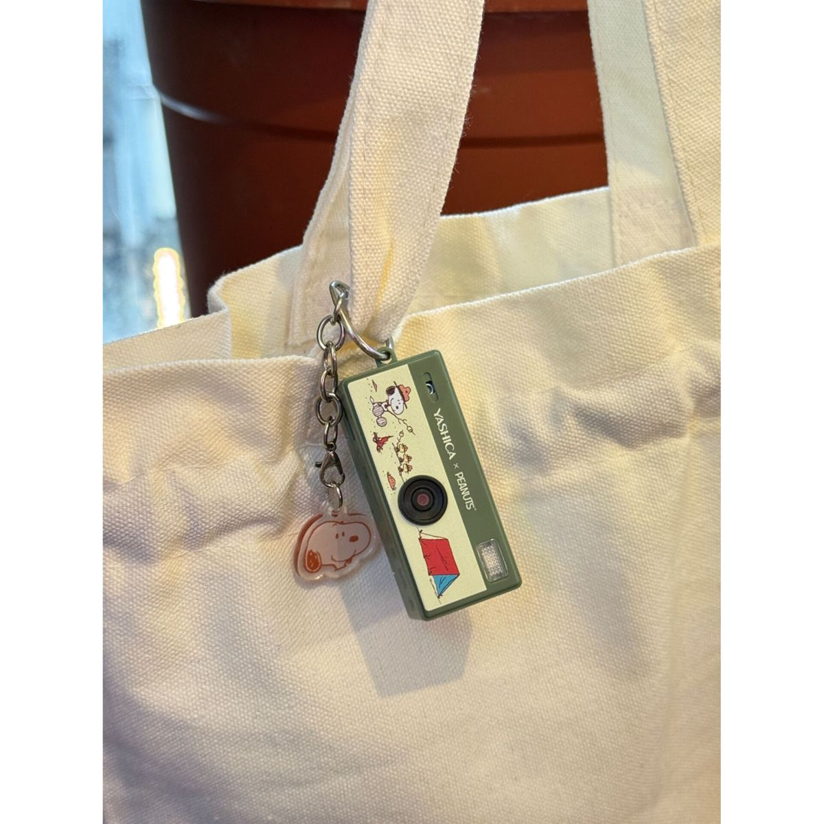 Ein olivgrüner Kamera-Schlüsselanhänger mit Snoopy-Motiv hängt am Riemen einer weißen Stofftasche.