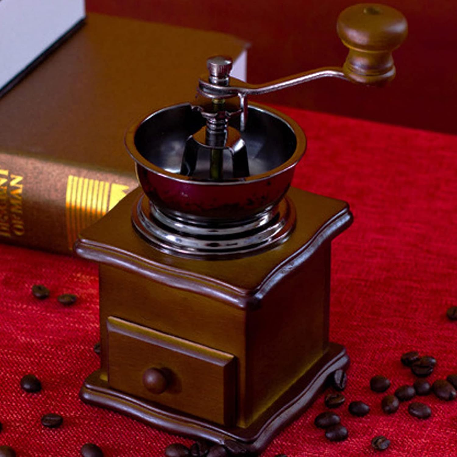 Eine alte Kaffeemühle aus Holz auf rotem Untergrund mit Kaffeebohnen und einem Buch im Hintergrund.