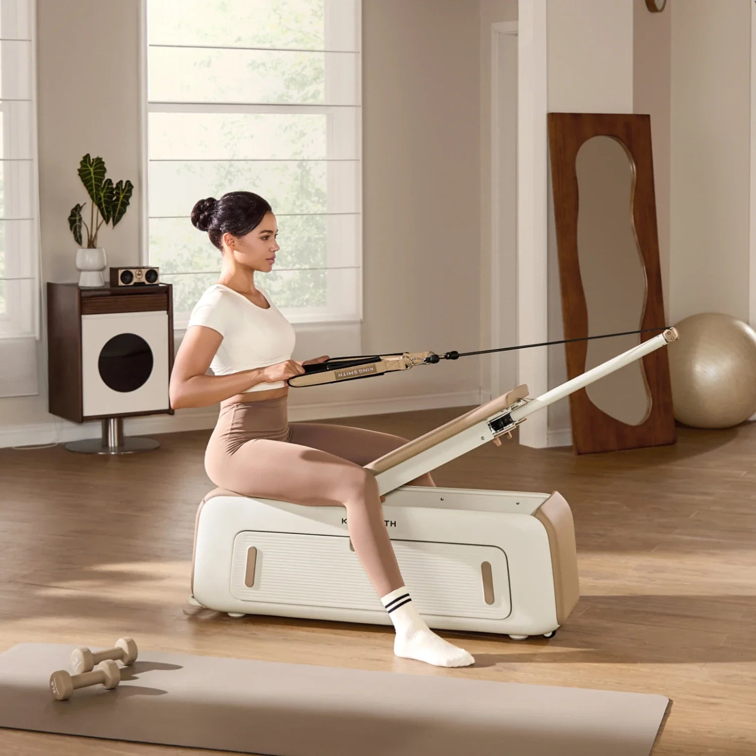 Una donna in abbigliamento sportivo siede su un attrezzo per pilates, tenendo delle cinghie, in una stanza luminosa.