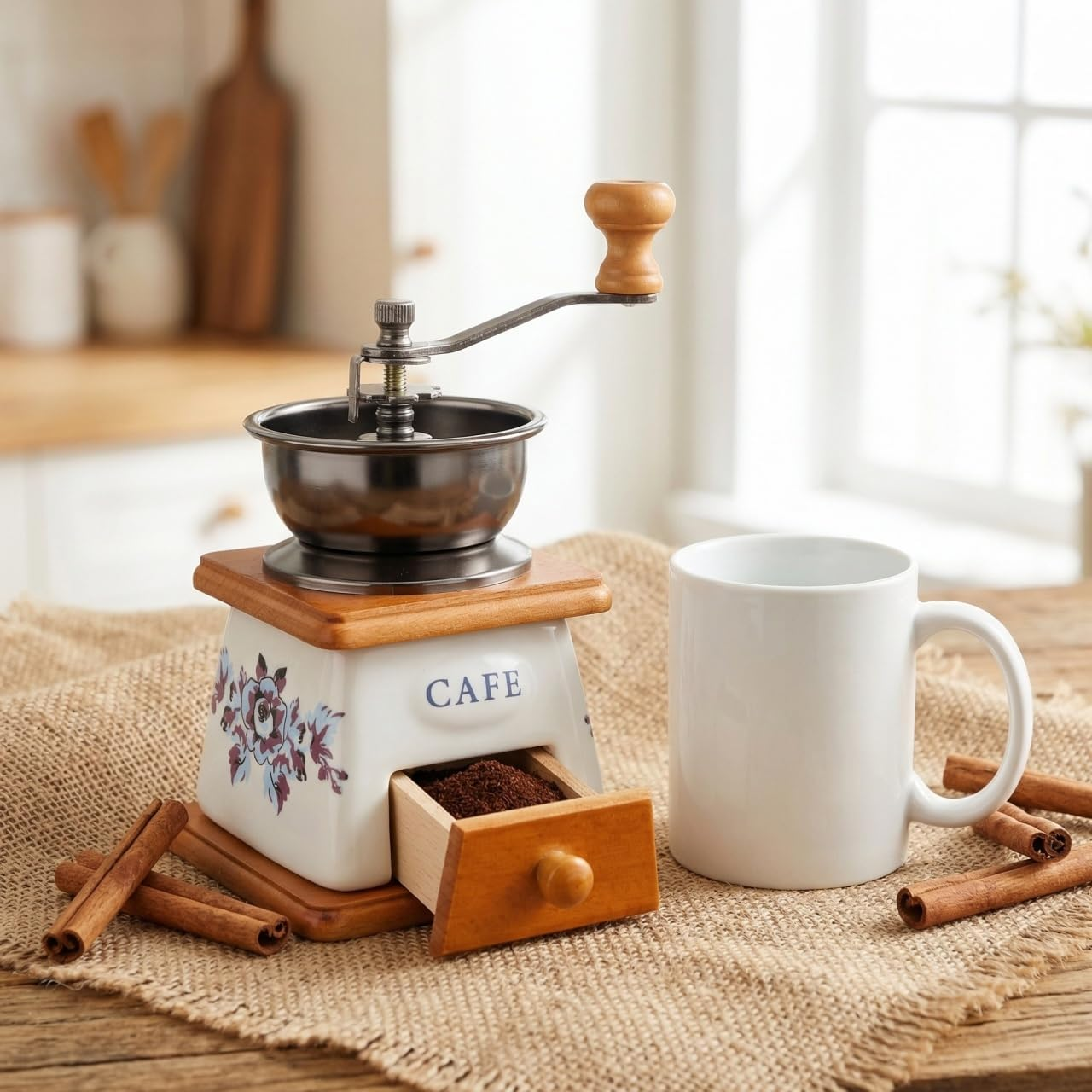 Eine Handkaffemühle mit offener Kaffeepulverschublade, eine weiße Tasse und Zimtstangen auf Jute.