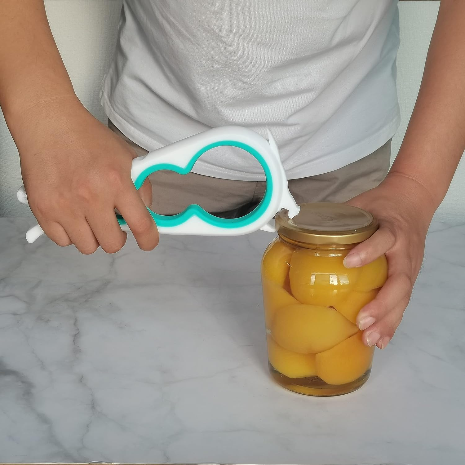 Eine Person benutzt einen weiß-türkisen Glasöffner bei einem Glas mit Pfirsichscheiben auf weißer Marmoroberfläche.