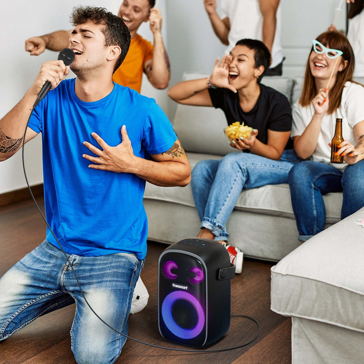 Ein Mann im blauen Shirt kniet und singt in ein Mikrofon, während Freunde Spaß haben und ein beleuchteter Partylautsprecher in der Nähe steht.
