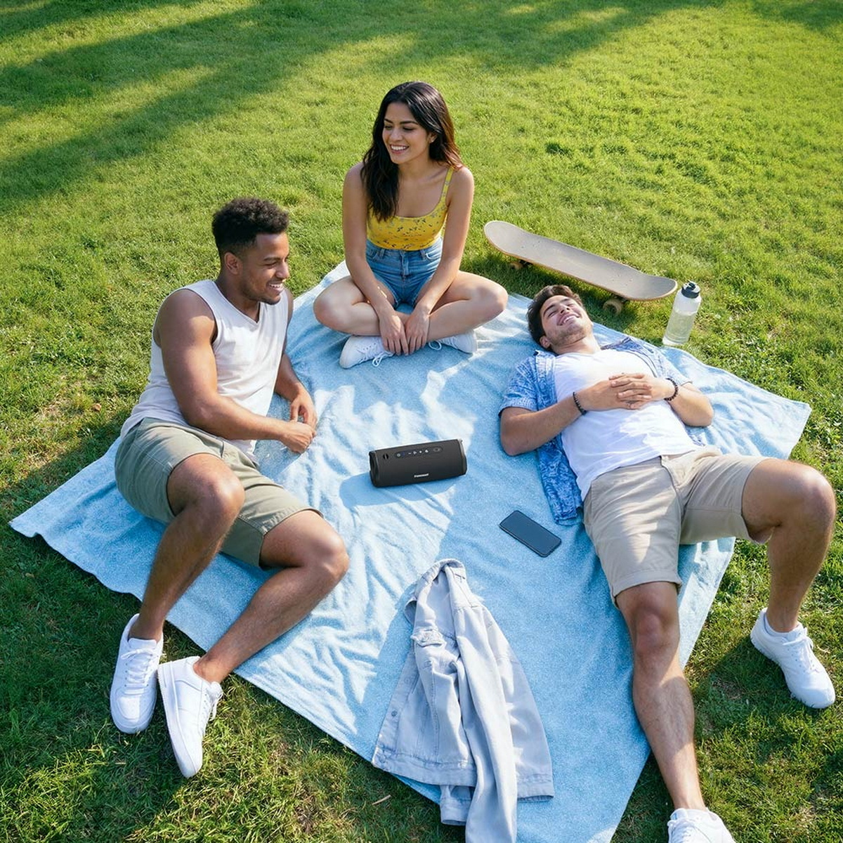Drei Freunde entspannen auf einer blauen Decke auf einer sonnigen grünen Wiese mit einem mobilen Lautsprecher.