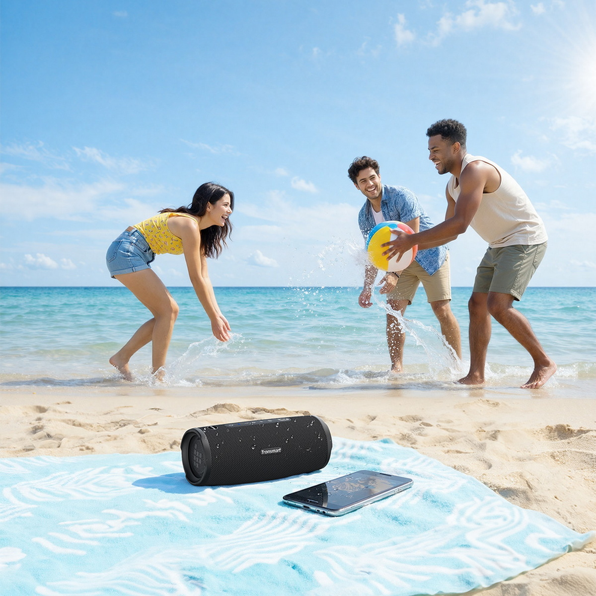 Drei Freunde spielen am Strand im flachen Wasser mit einem Ball, während ein schwarzer Lautsprecher im Vordergrund auf einem blau-weißen Handtuch liegt.