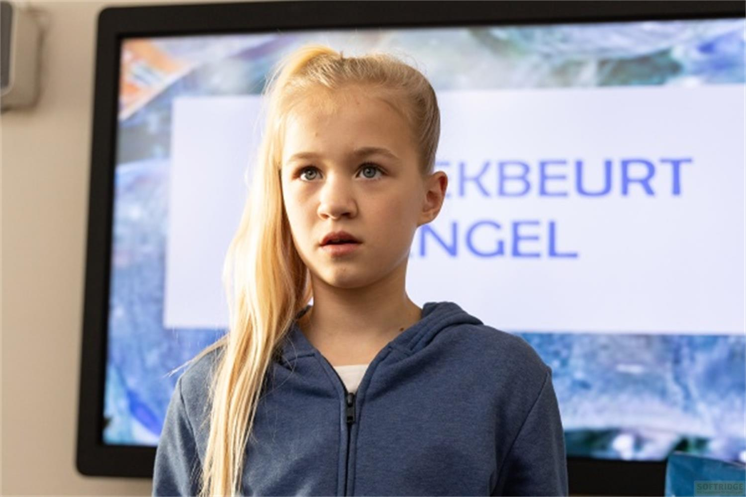 Ein junges blondes Mädchen in einem blauen Kapuzenpullover vor einem Bildschirm mit Text.