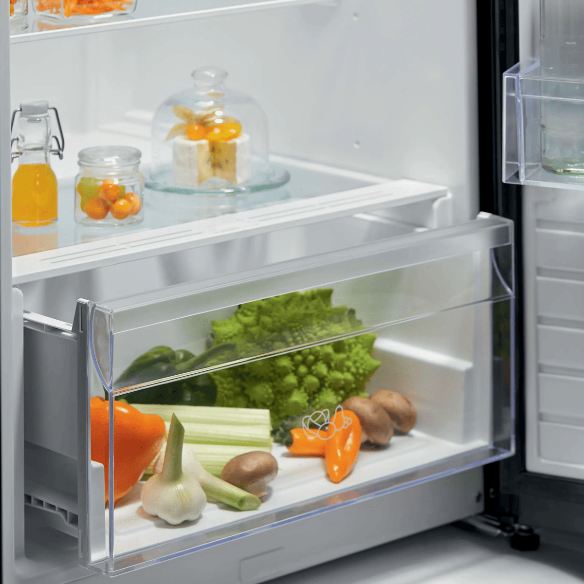 Refrigerador abierto con estantes de vidrio que muestran quesos, frascos y un cajón inferior con verduras.