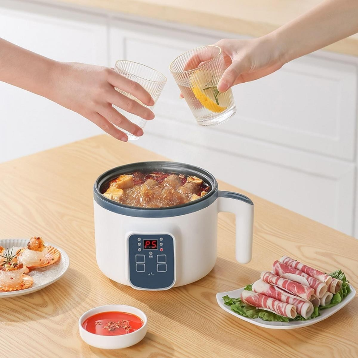 Zwei Personen halten Gläser über einem kleinen weißen Kocher mit Suppe auf einem Holztisch, daneben Fleisch und Sauce.