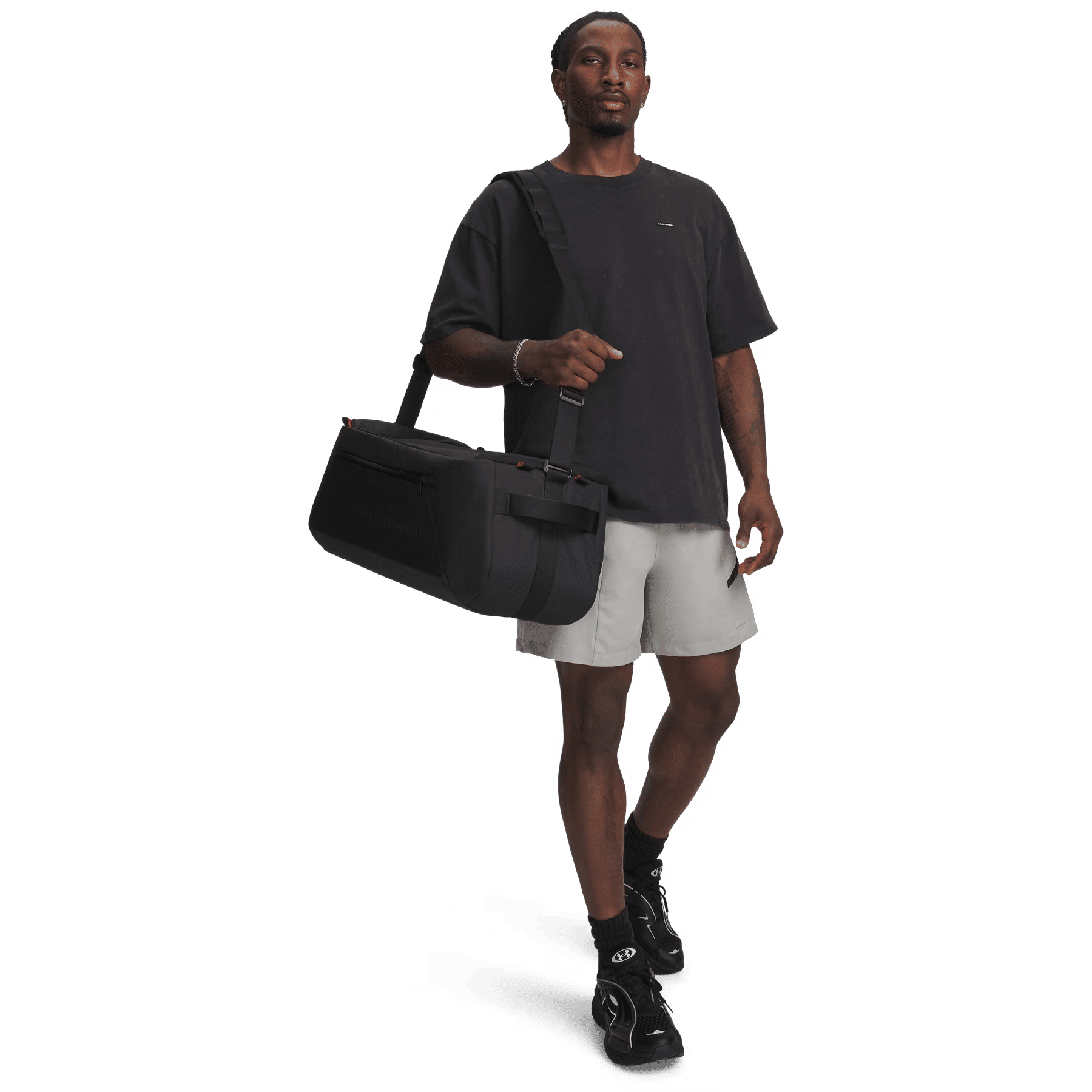 Ein Mann im schwarzen T-Shirt und grauen Shorts hält eine schwarze Sporttasche und geht vor schwarzem Hintergrund.