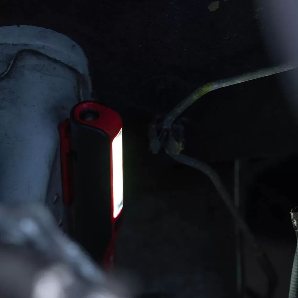 Kompaktowa czerwono-czarna lampa robocza oświetla ciemną komorę silnika.