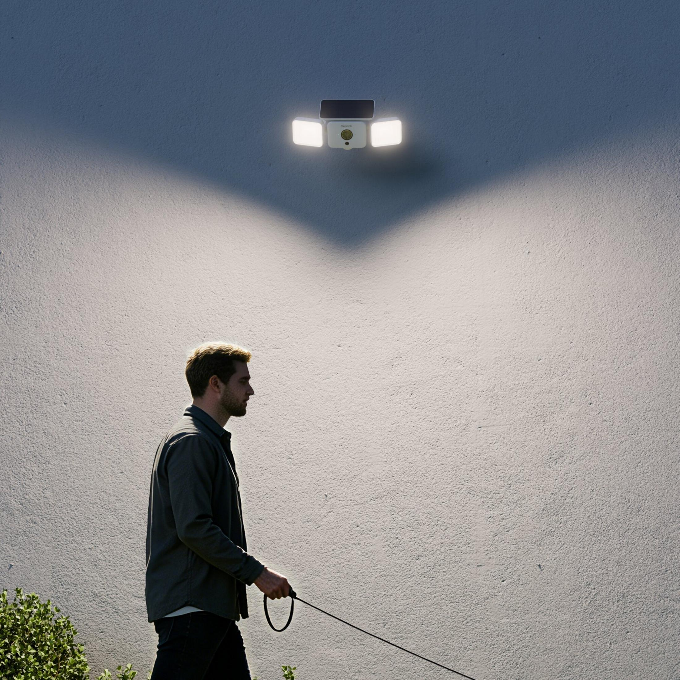 Ein Mann geht im hellen Licht einer an einer grauen Wand montierten Sicherheitskamera mit LED-Flutlicht.