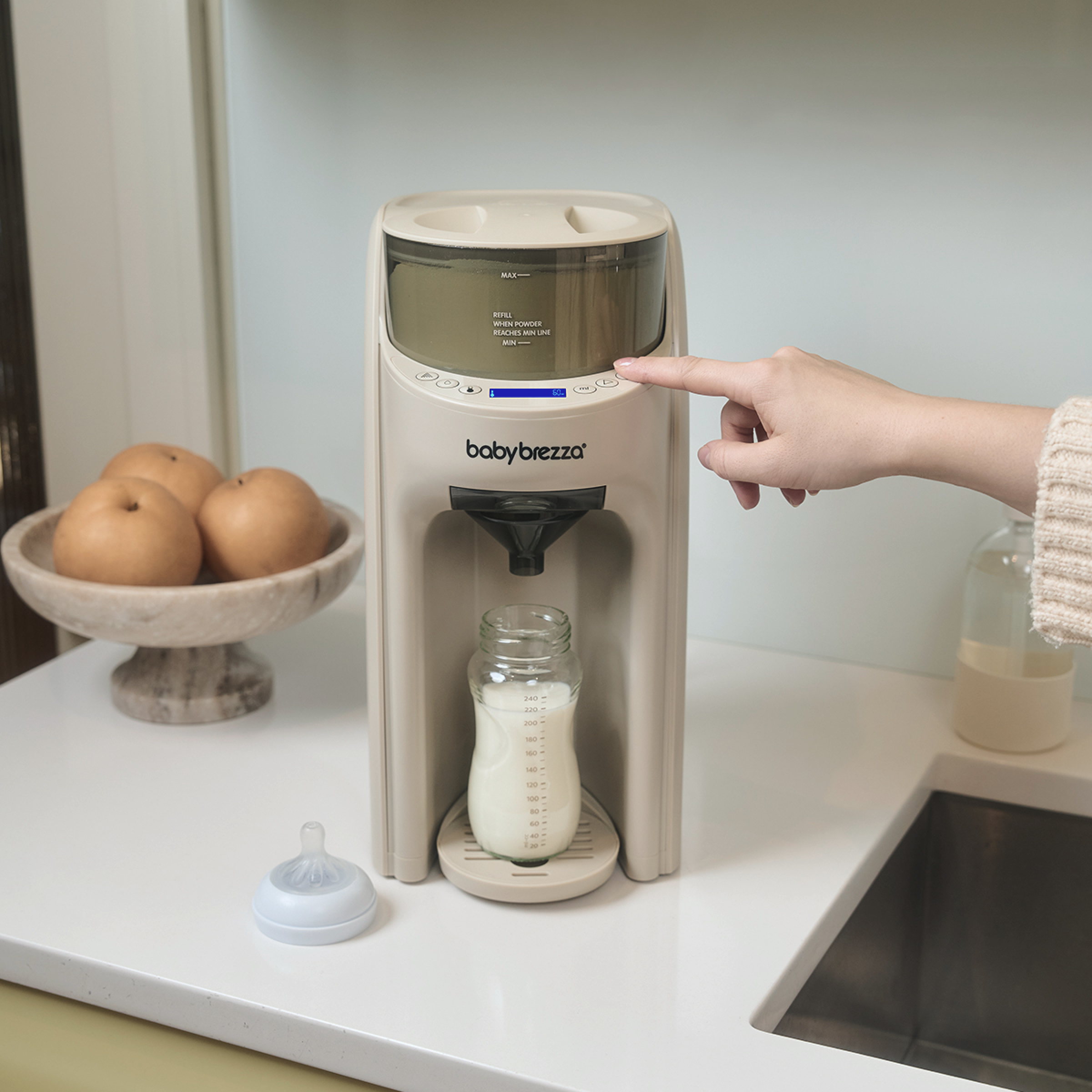 Eine Hand drückt den Startknopf einer Babybrezza-Maschine auf einer Küchentheke mit einer Milchflasche.