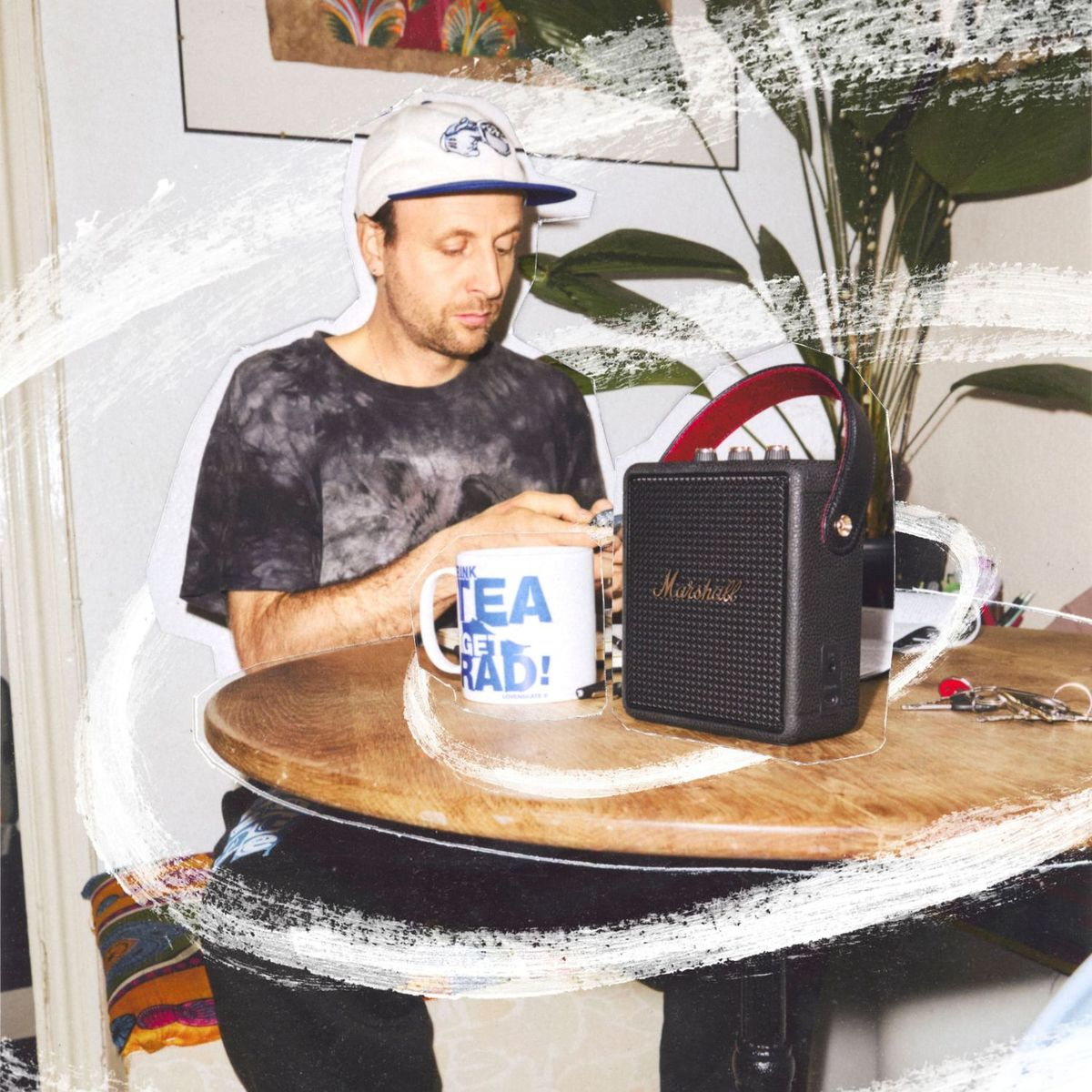 Ein Mann mit Mütze und Batikshirt sitzt an einem Holztisch mit weißer Tasse und einem kleinen schwarzen Marshall-Lautsprecher.