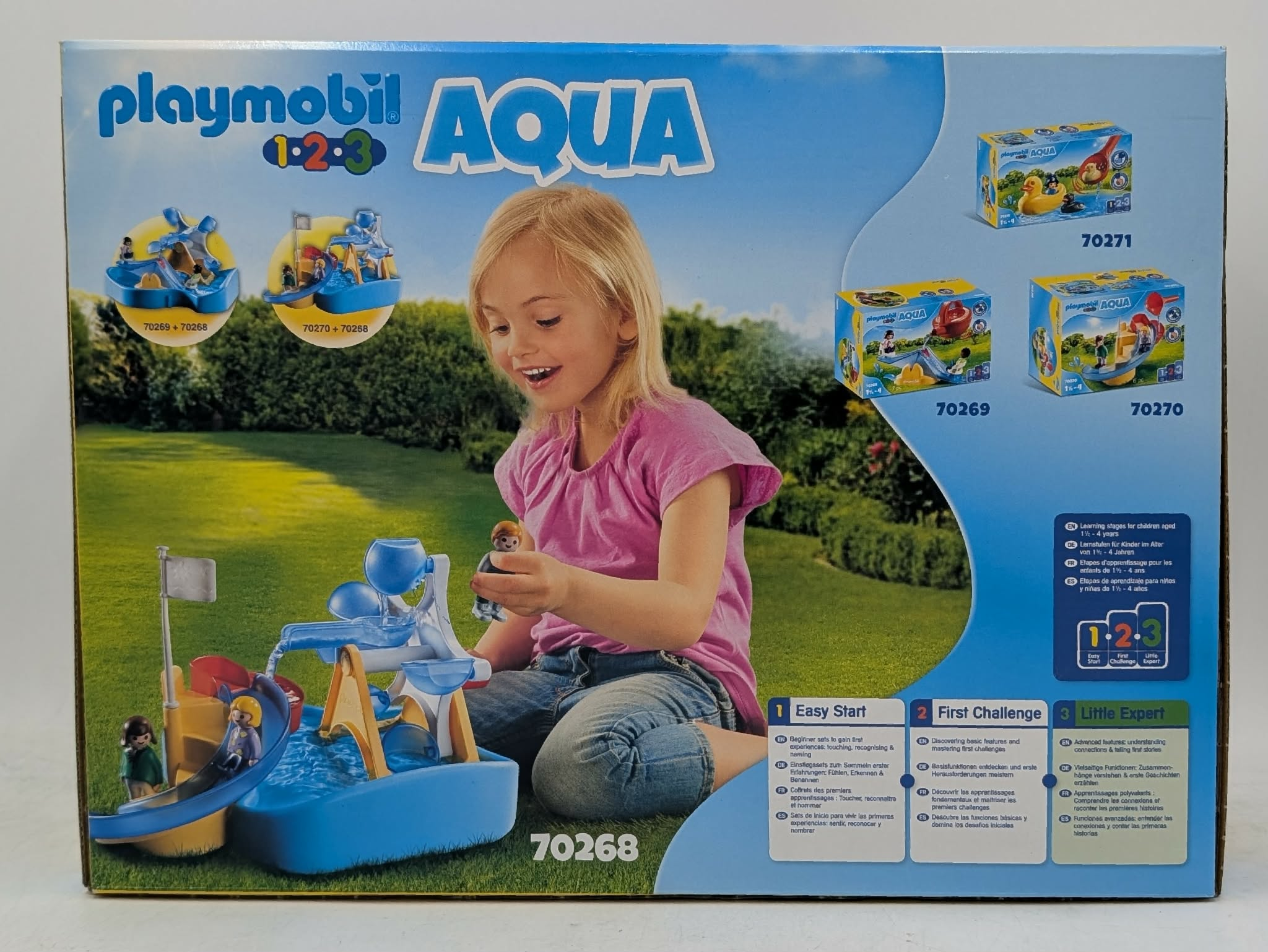 Ein blondes Mädchen sitzt im Garten vor einer Spielzeugschachtel von Playmobil Aqua und spielt mit dem blauen Wasserspielzeug.