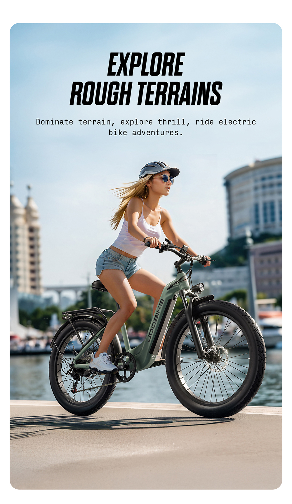Una donna bionda guida una bicicletta elettrica verde su una passeggiata con edifici e acqua.