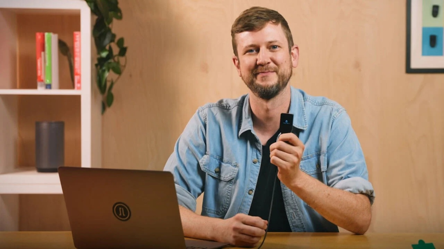 Ein lächelnder Mann sitzt im Büro vor einem Laptop und hält ein kleines technisches Gerät.