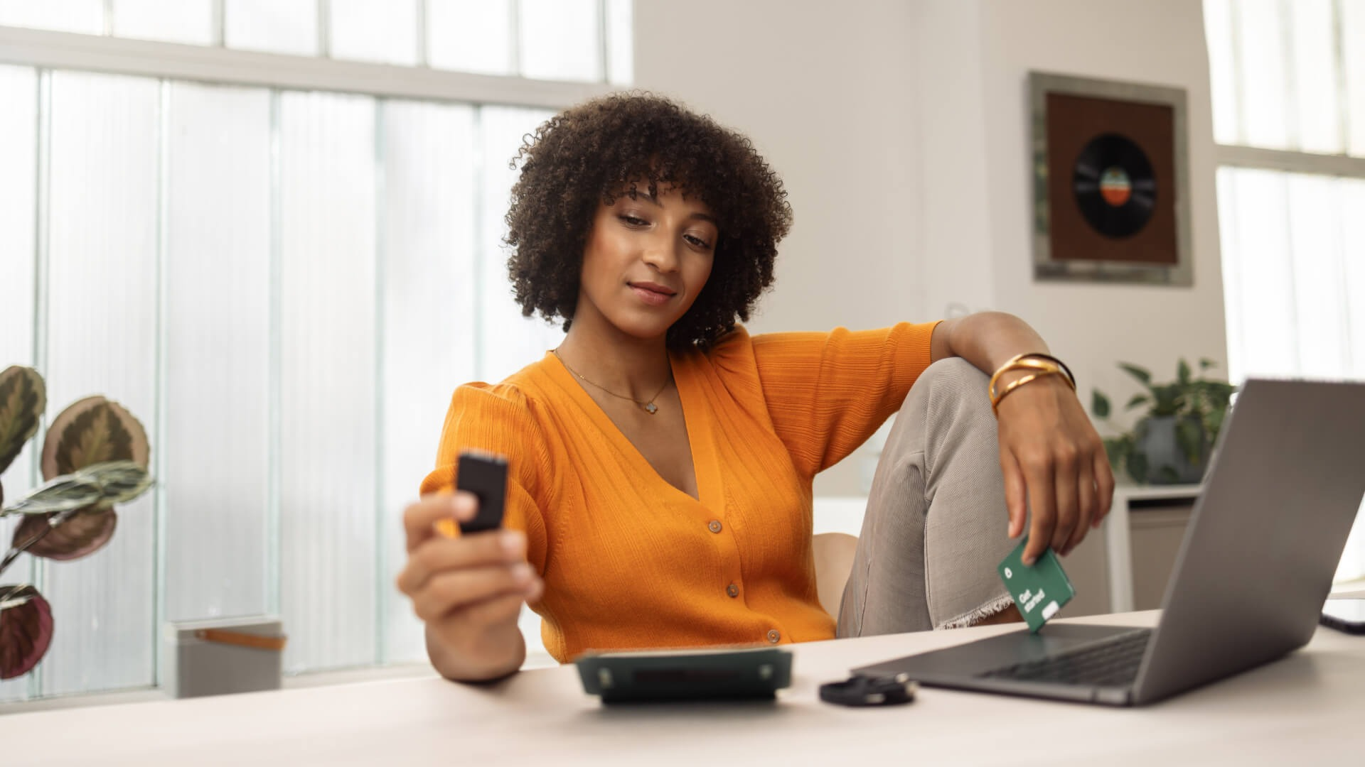 Eine Frau mit Locken in einem orangen Pullover sitzt an einem Tisch mit einem Laptop und hält eine Karte.