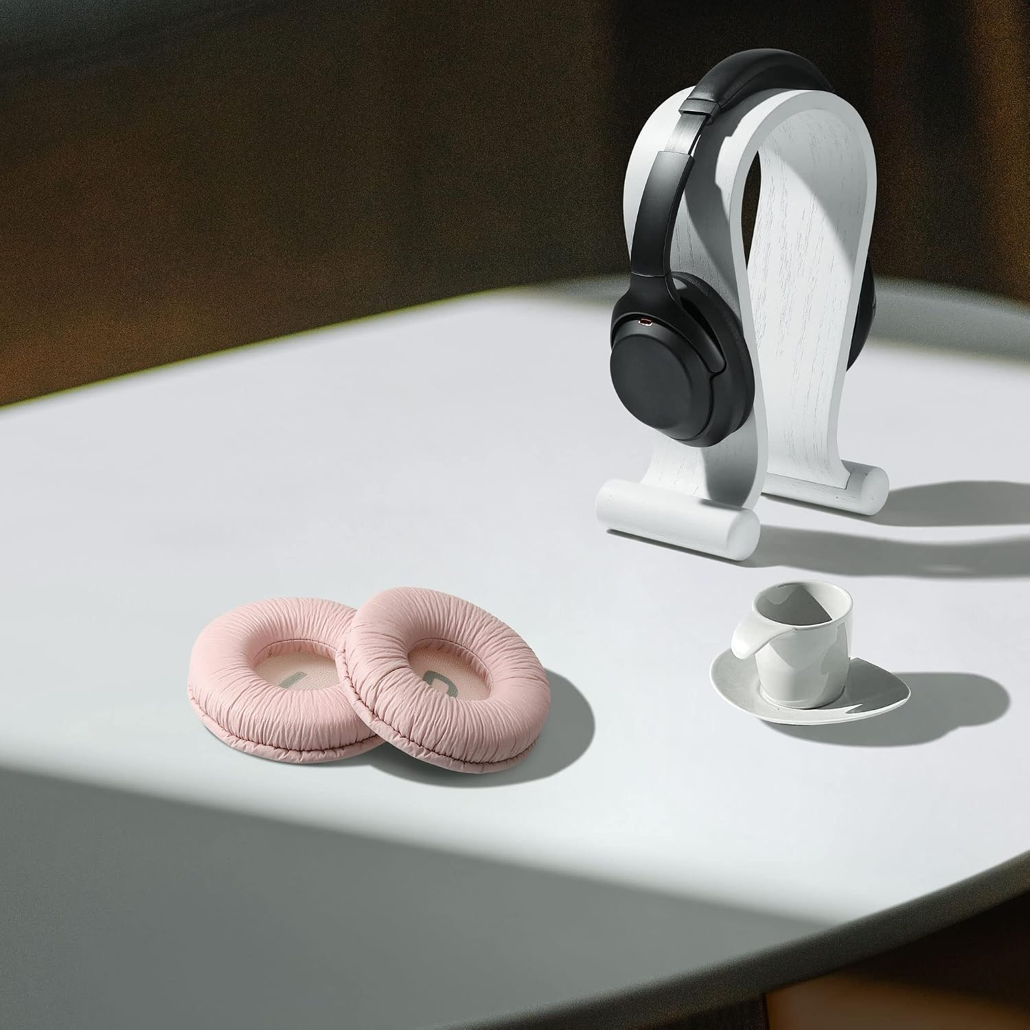 Schwarze Kopfhörer auf weißem Ständer, zwei rosa Ohrpolster und eine kleine weiße Kaffeetasse auf einem Tisch.