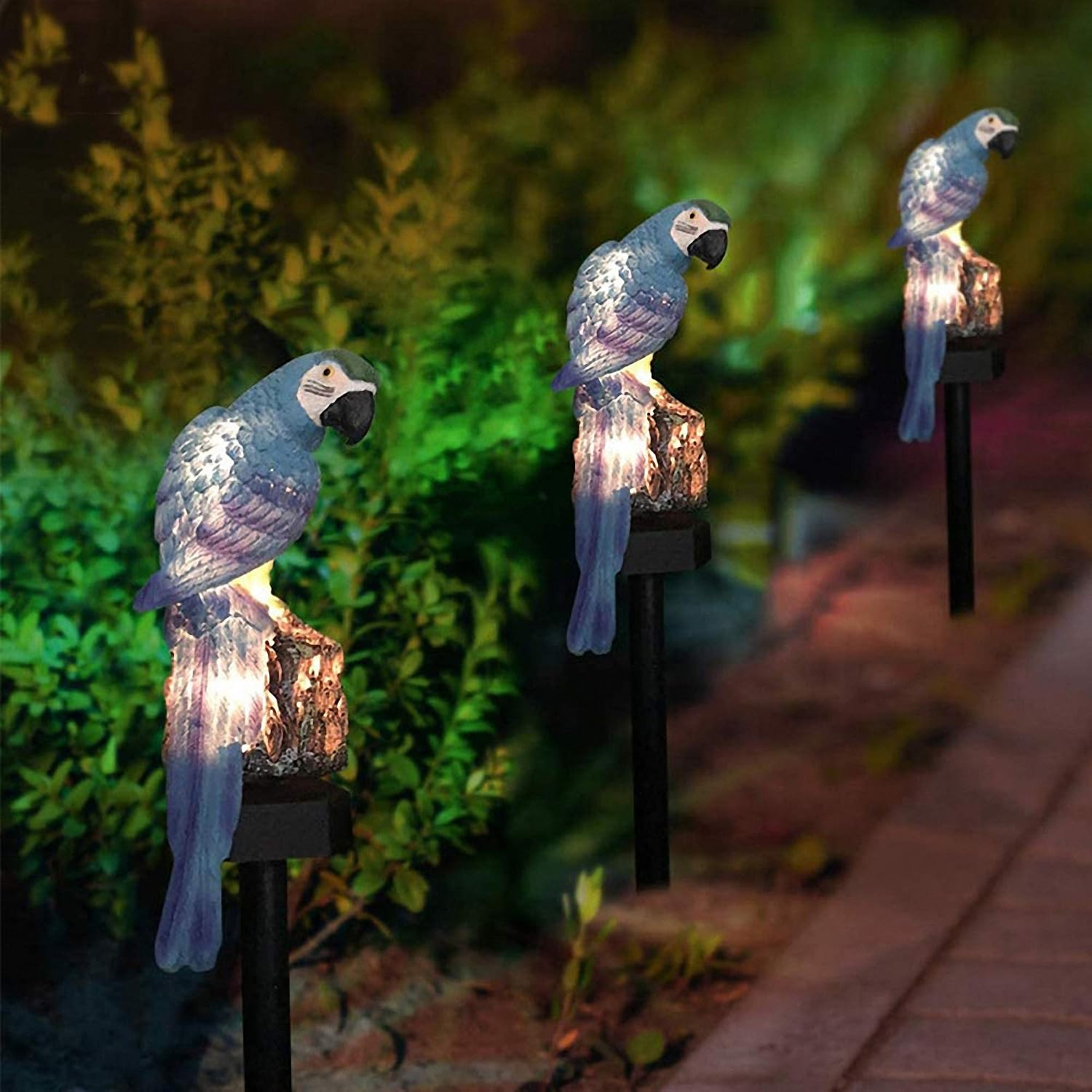 Drei blaue Papageien-Gartenleuchten stehen nachts beleuchtet entlang eines Gartenweges.