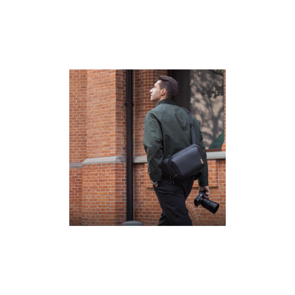 Un uomo in giacca verde cammina davanti a un muro di mattoni, con borsa nera e fotocamera.
