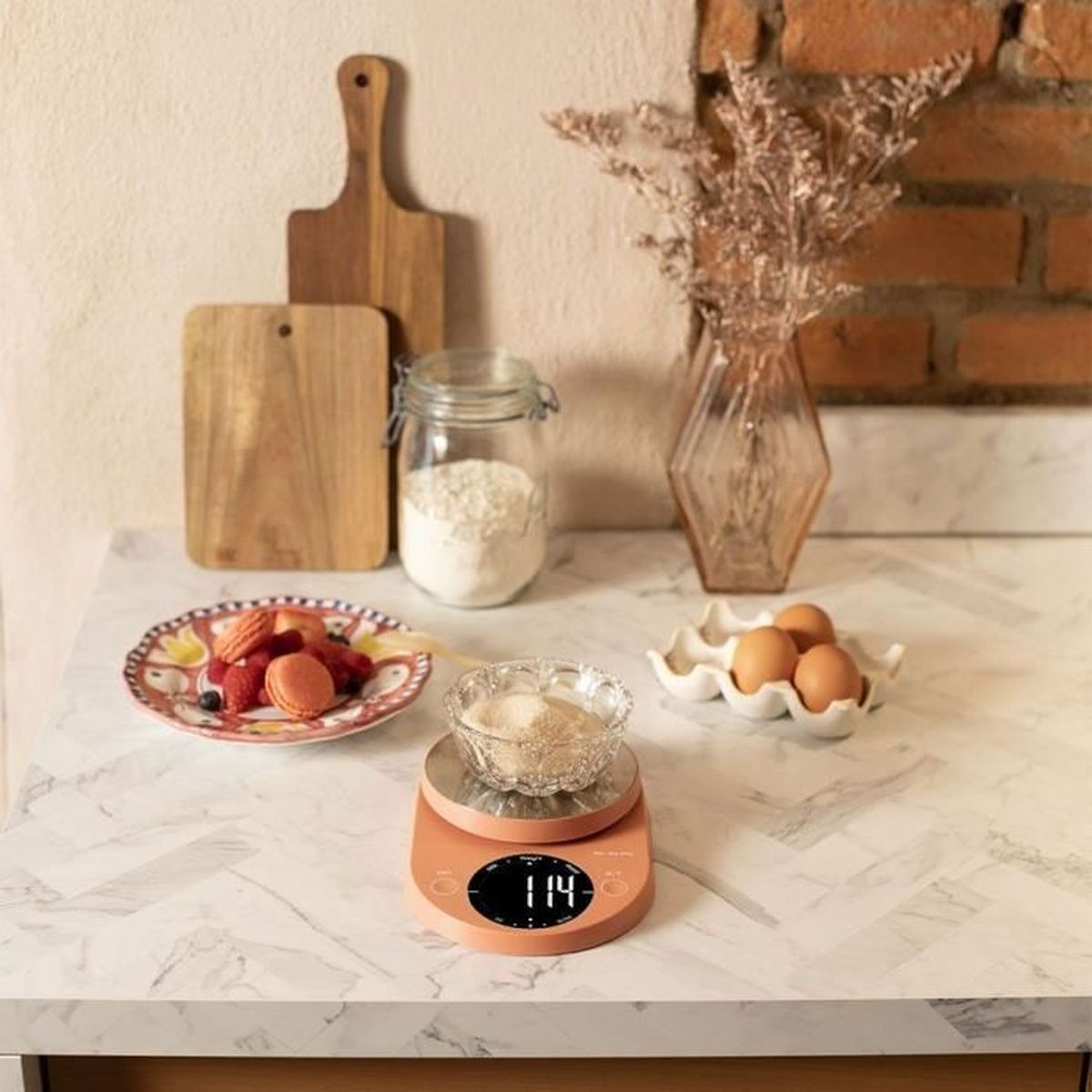 Küchenwaage auf weißer Marmorarbeitsplatte mit Macarons, Eiern und Glasbehältern.