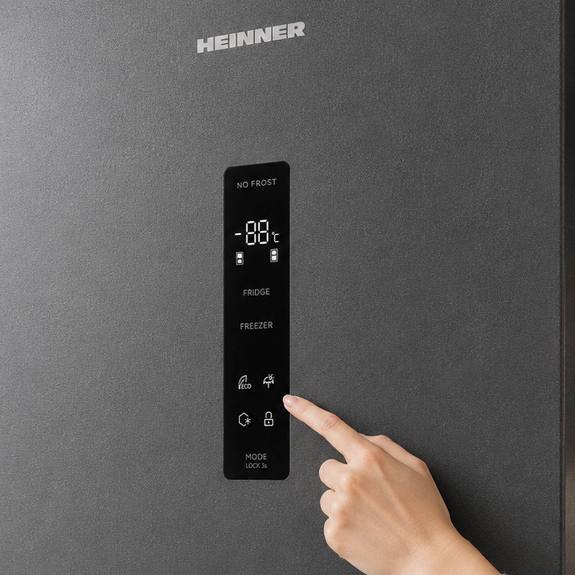 Une main touche l'écran tactile numérique d'un réfrigérateur gris Heinner.