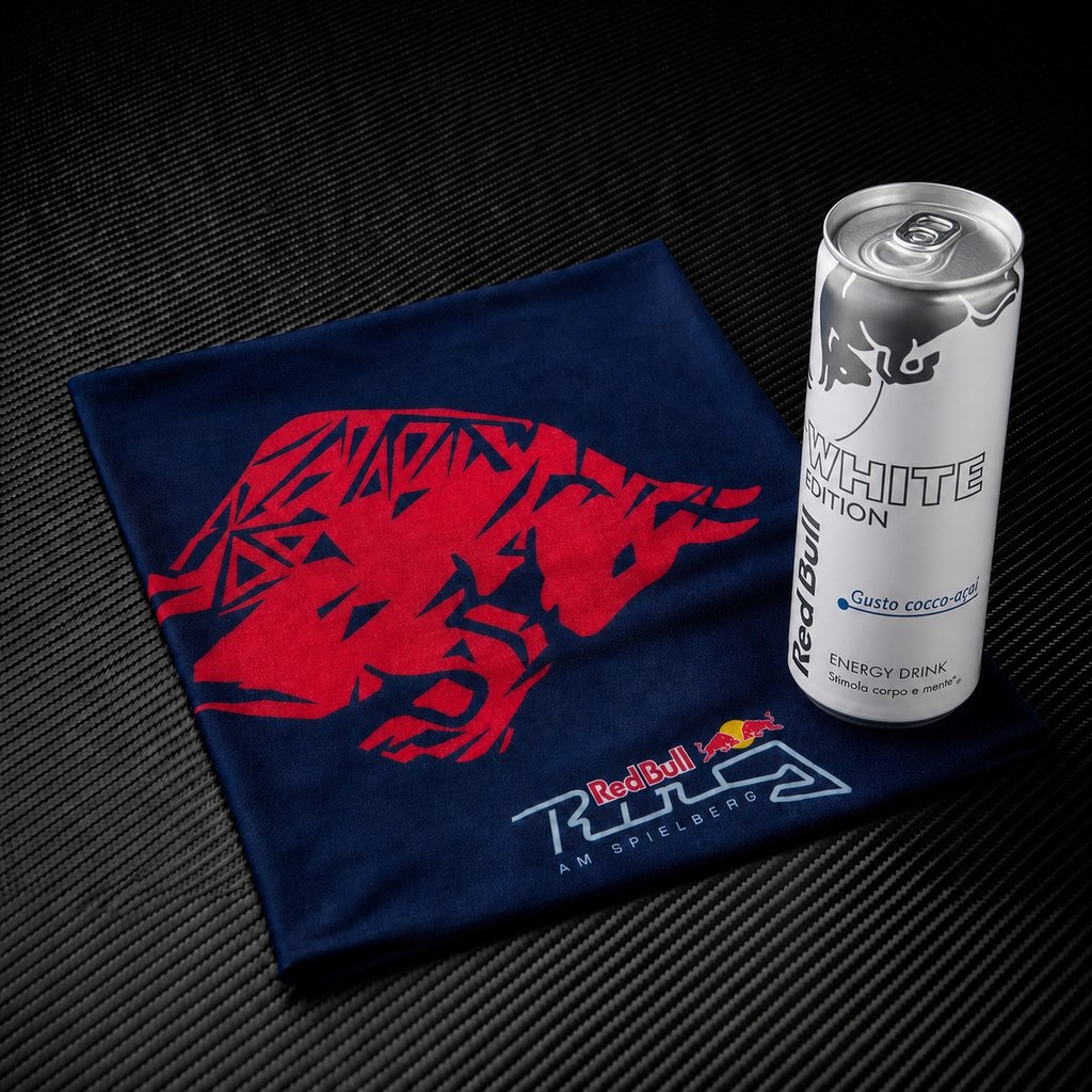 Una lattina bianca di Red Bull accanto a un panno blu scuro con logo toro rosso su sfondo nero.