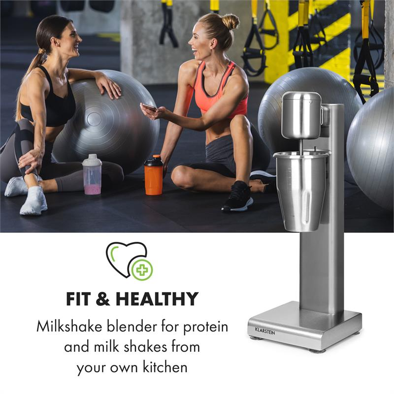 Silberner Milchshake-Mixer vor einem Fitnessstudio mit zwei Frauen in Sportkleidung.