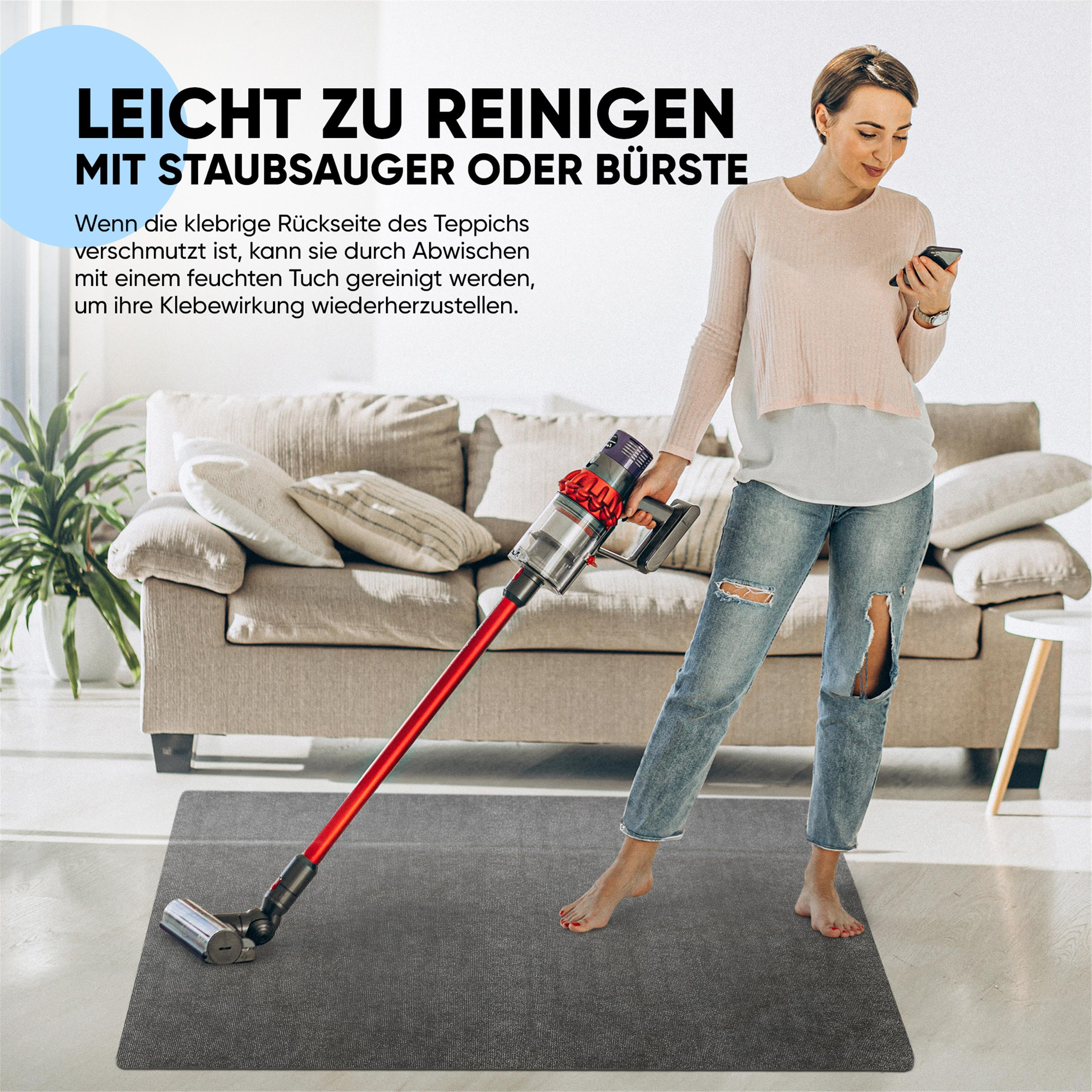 Eine Frau staubsaugt einen grauen Teppich und hält ihr Smartphone, mit Sofa im Hintergrund.
