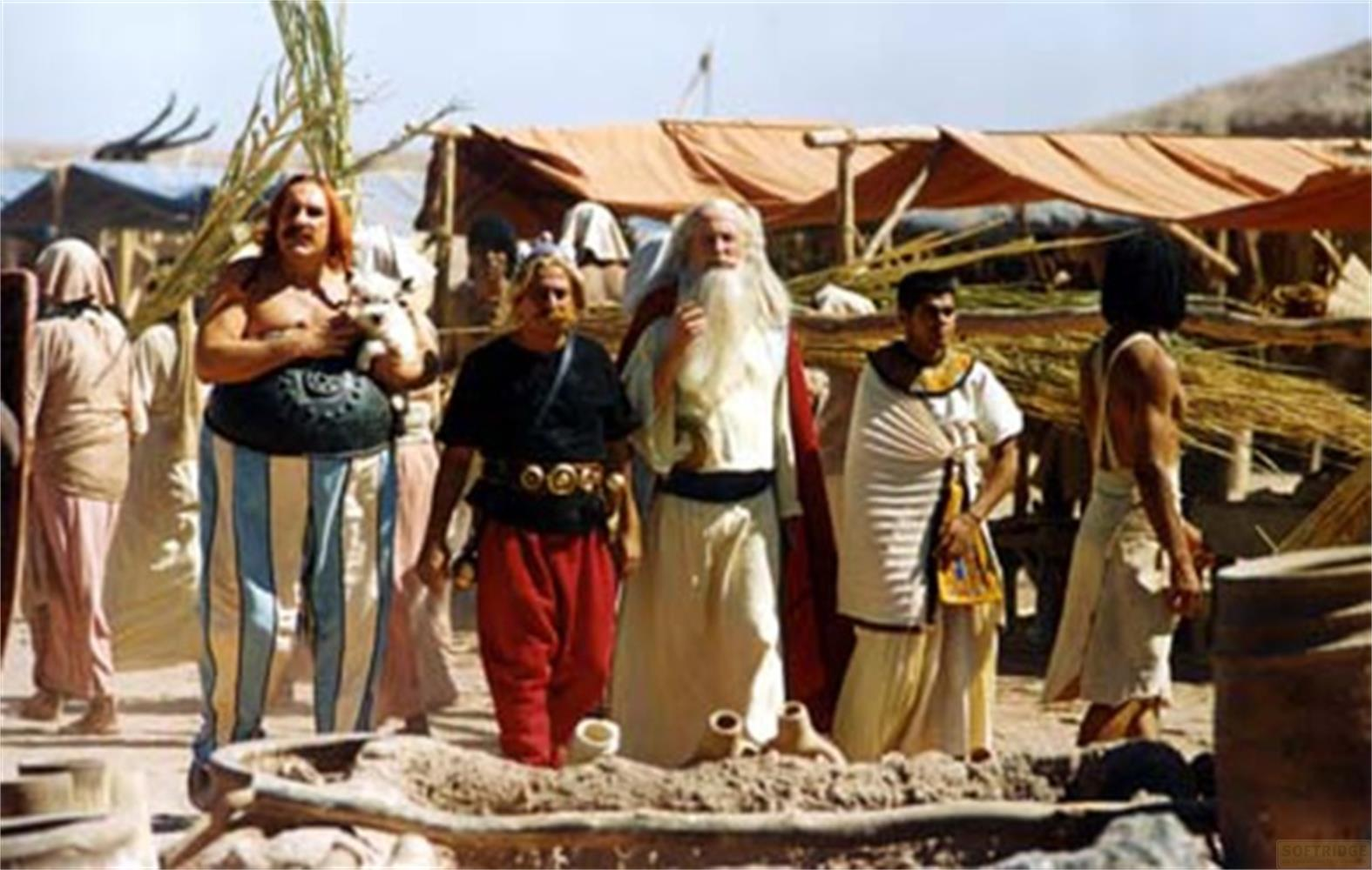 Obelix, Asterix und Miraculix gehen mit anderen Figuren unter Zeltdächern über einen geschäftigen Markt.