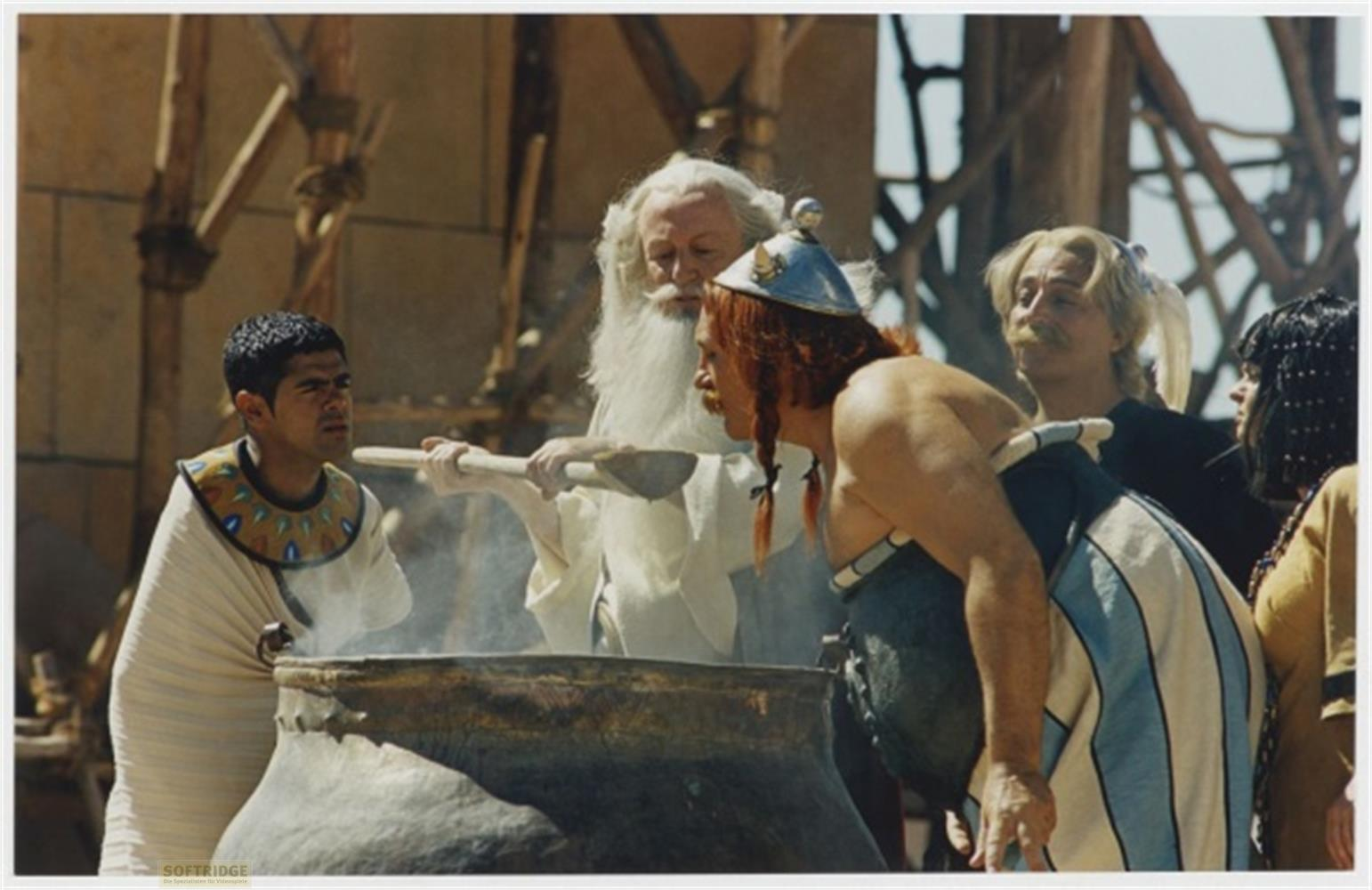 Ein Druide mit weißem Haar rührt mit einem Holzlöffel in einem großen Topf, umgeben von Figuren in antiken Kostümen.