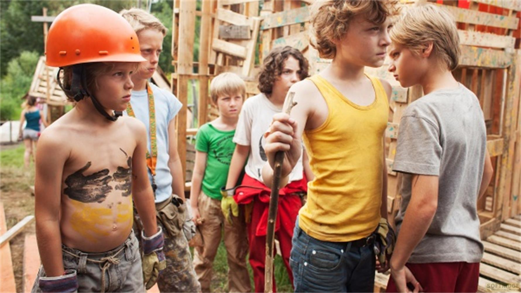 Eine Gruppe von Kindern steht im Freien vor einer Holzkonstruktion, wobei sich zwei Jungen direkt gegenüberstehen.