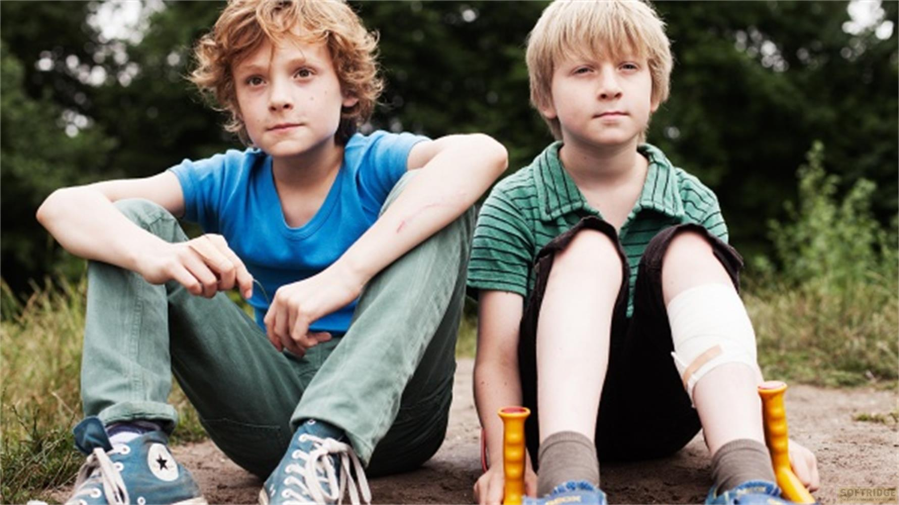 Zwei Jungen mit blonden Haaren sitzen im Freien auf dem Boden, einer trägt ein blaues T-Shirt und der andere ein grünes Hemd.