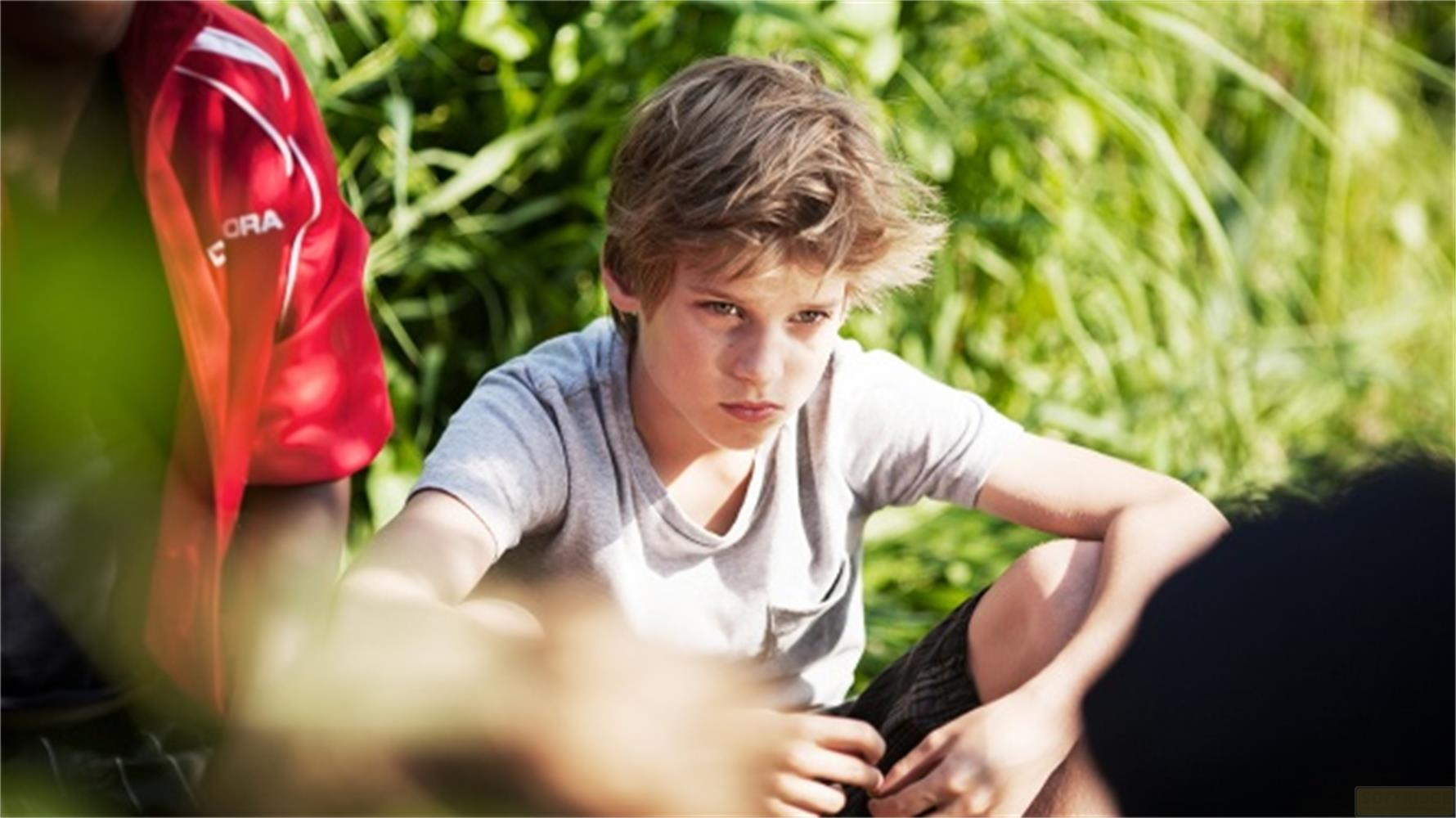 Ein Junge in einem grauen T-Shirt sitzt draußen im Gras mit anderen Personen.